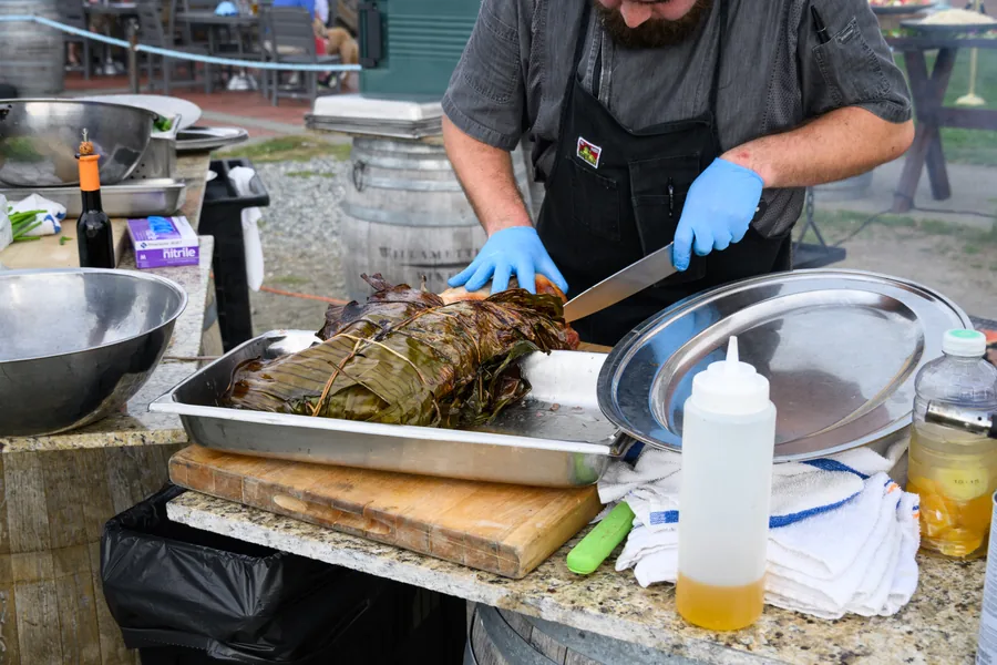 Chef Blake in an apron carving into a leaf-wrapped roast on a cutting board at an outdoor prep station