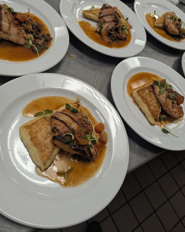 A line of identical plates of sliced chicken over polenta cakes with pan sauce and microgreens, ready for service
