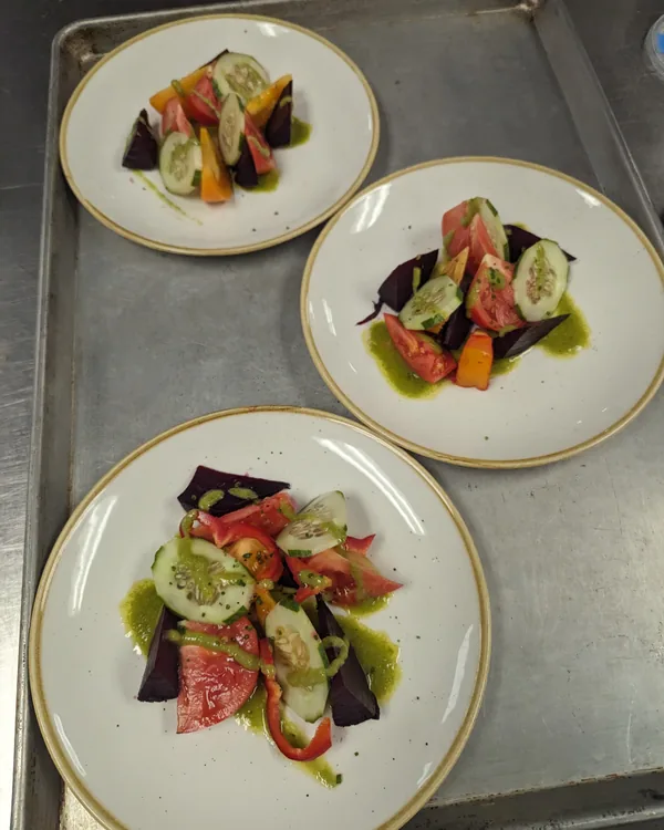 Three matching plates of heirloom tomato and roasted beet salad with herb oil, lined up on a steel tray
