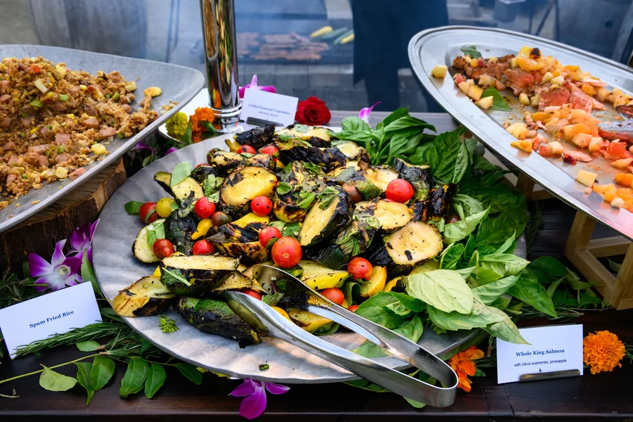 Large oval platters of grilled vegetable salad, orzo and sliced meats laid out with orchids for an event buffet