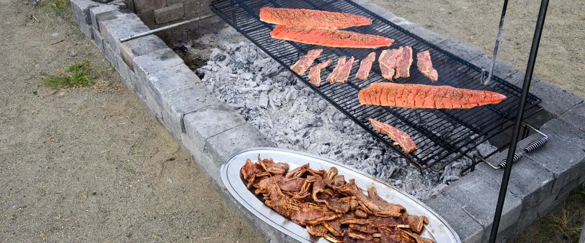 An outdoor fire pit grill loaded with salmon and steaks over coals, with a large oval platter of finished meat resting beside it