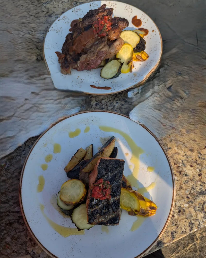 Two plates of seared steak with herb salsa, blistered squash and roasted potatoes, plated outdoors on stone